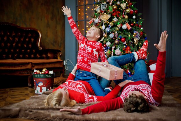 Ritratto della famiglia di Natale in maglioni tradizionali rossi nel salone, nei genitori e nel bambino di festa domestica con il contenitore di regalo.