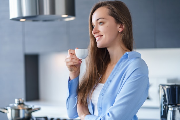 Ritratto della femmina attraente felice che beve caffè aromatico fresco caldo dopo la preparazione del caffè facendo uso della macchinetta del caffè nella cucina a casa