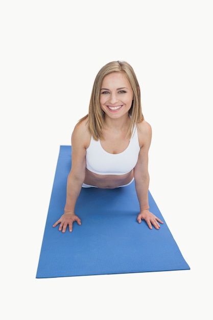 Ritratto della giovane donna felice che fa i piegamenti sulle braccia sulla stuoia di esercizio sopra fondo bianco