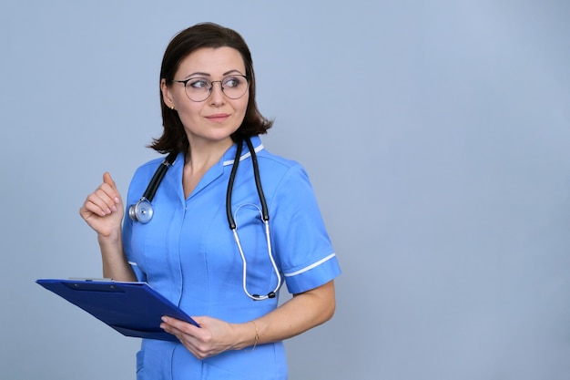 Ritratto della lavagna per appunti matura della tenuta della donna dell'infermiera. Femmina in uniforme blu con stetoscopio su sfondo grigio, copia spazio