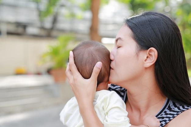 Ritratto della madre asiatica che porta e che bacia il suo neonato infantile all&#39;aperto.