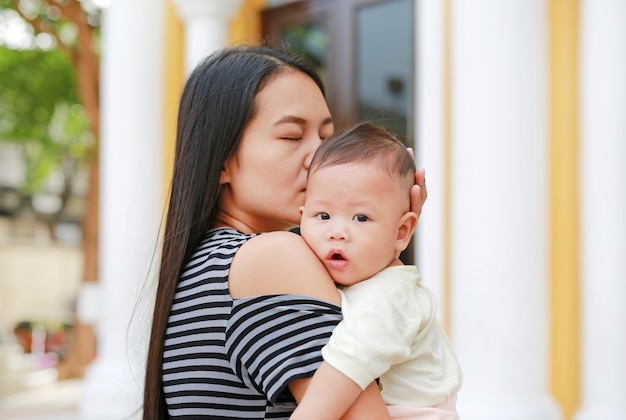 Ritratto della madre asiatica che porta e che bacia il suo neonato infantile all&#39;aperto.