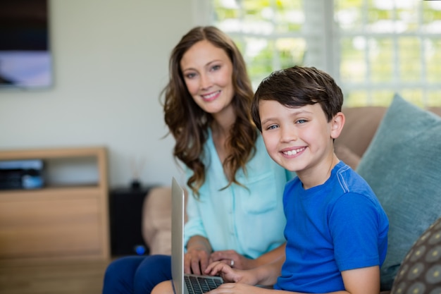 Ritratto della madre e del figlio sorridenti che si siedono sul sofà per mezzo del computer portatile