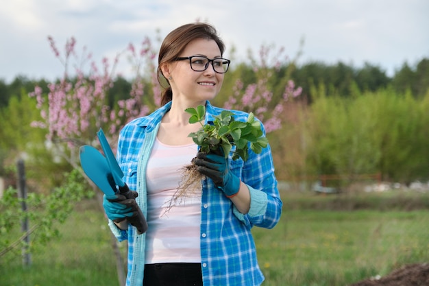 Ritratto della primavera della donna matura in giardino con gli strumenti, cespugli di fragola.