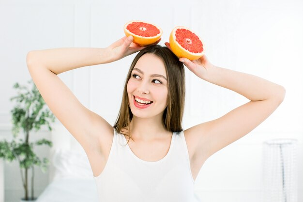 Ritratto della ragazza abbastanza positiva attraente allegra che ha due pezzi di pompelmo in sue mani al di sopra sui precedenti leggeri. Concetto di cibo sano. Vitamine e minerali.