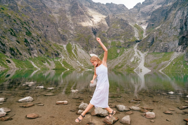 Ritratto della ragazza bionda che equilibra alla pietra in lago fra le montagne.