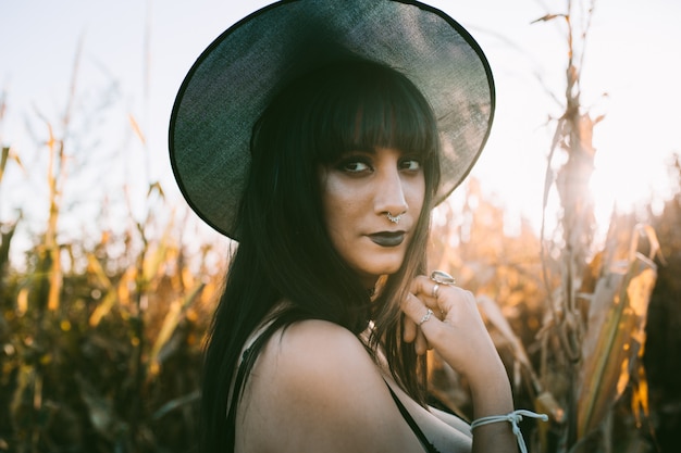 Ritratto della ragazza della strega del costume di Halloween in un campo di mais al tramonto. Bella giovane donna seria in cappello delle streghe con lunghi capelli neri e labbra scure