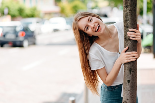Ritratto della ragazza felice di divertimento con l'ampio sorriso che sta e che abbraccia un albero all'aperto