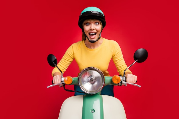 Ritratto della ragazza felice pazza adorabile che guida il fronte eccitato del ciclomotore sulla parete rossa