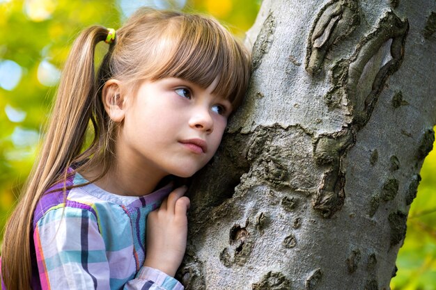 Ritratto della ragazza graziosa del bambino che si appoggia ad un tronco di albero nel rilassamento del parco di autunno. Bambino femminile sveglio che gode del tempo caldo di autunno in foresta.