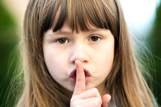 Ritratto della ragazza graziosa del bambino con gli occhi grigi e il dito lungo del punto di tenuta dei capelli biondi lunghi sulle sue labbra con silenzio sospiro. Bambino femminile sveglio il giorno di estate caldo fuori.