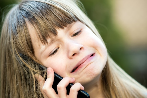 Ritratto della ragazza sollecitata del bambino con capelli lunghi che parla sul telefono cellulare. Piccolo bambino femminile che comunica facendo uso dello smartphone. Concetto di comunicazione dei bambini.