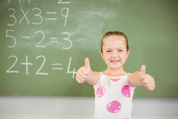 Ritratto della scolara sorridente che mostra i pollici su nell'aula