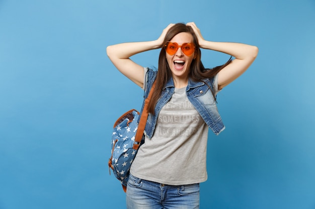 Ritratto della studentessa allegra felice emozionante con lo zaino in vetri del cuore arancio dei vestiti del denim che si aggrappano alla testa isolata su fondo blu. Istruzione al liceo. Copia spazio per la pubblicità.