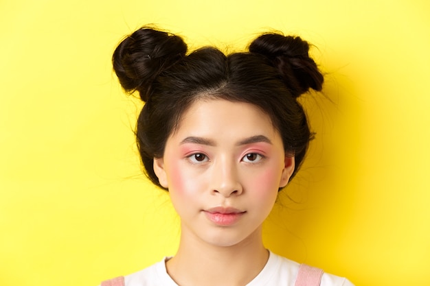 Ritratto della testa della ragazza asiatica alla moda con trucco luminoso e hairbuns, che guarda l'obbiettivo, in piedi sul giallo.