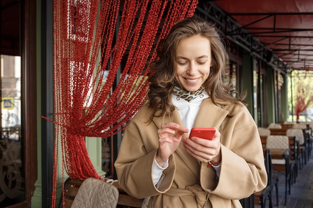 Ritratto della via di giovane donna sorridente che porta cappotto beige facendo uso del suo Smart Phone su una veranda del caffè