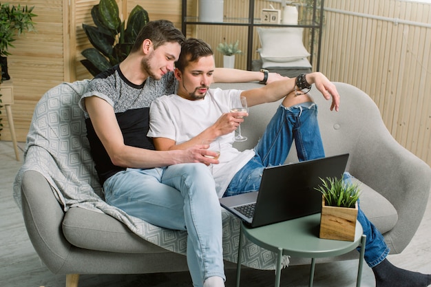 Ritratto delle coppie gay che si rilassano con il vino bianco sul sofà e che guardano il computer.