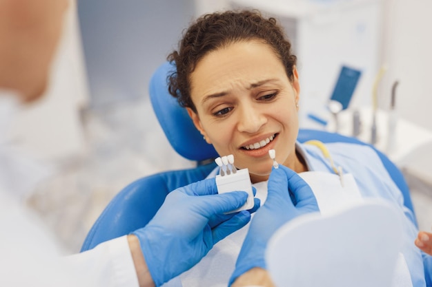 Ritratto delle mani del dentista in guanti di gomma che tengono la tavolozza dei colori dei denti con una paziente scontenta sulla poltrona del dentista