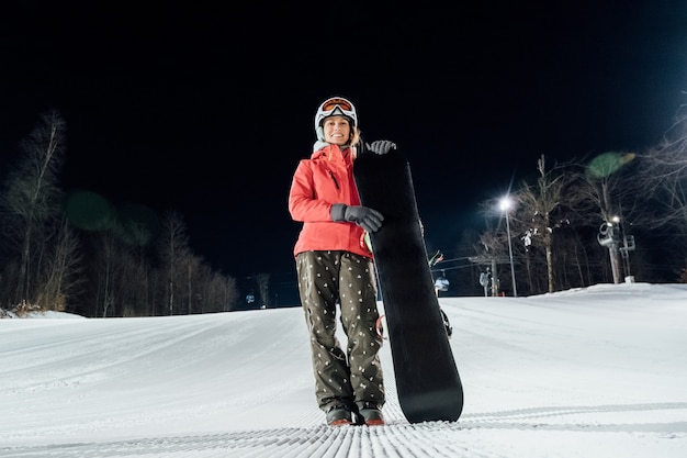 Ritratto dello snowboarder femminile al pendio di sera. Concetto di sport invernali