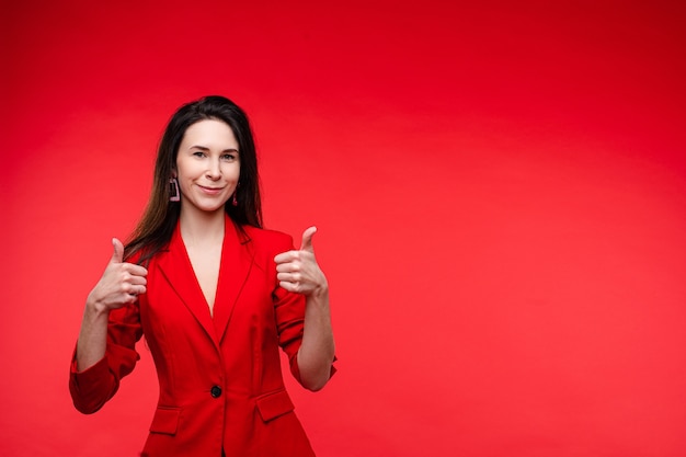 Ritratto dello studio di riserva di una donna adulta dai capelli scuri attraente in giacca rossa elegante che tiene i pollici in su e sorride alla macchina fotografica. Isolato su sfondo rosso. Copia spazio. Gesto di approvazione.