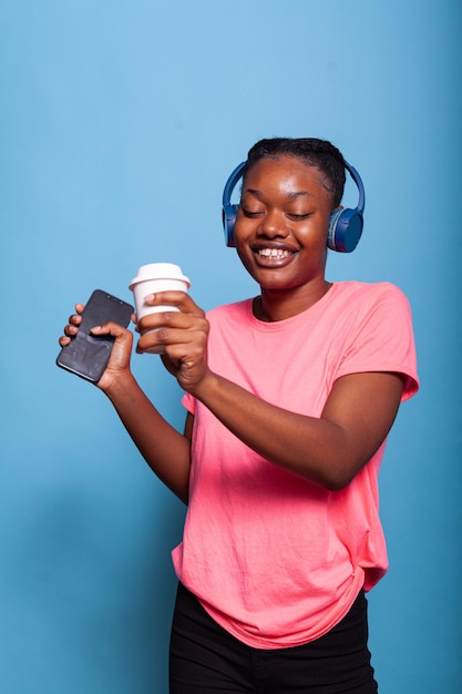 Ritratto di adolescente afroamericano con le cuffie che si godono la musica che tiene smartphone e caffè divertendosi in studio con sfondo blu. Studente che fa la mossa di danza. Audio e ritmo