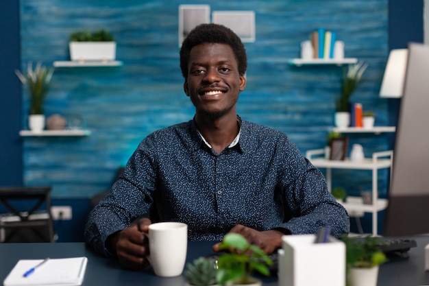 Ritratto di adolescente sorridente che tiene una tazza di caffè mentre studia alla lezione di gestione utilizzando la classe universitaria online durante il blocco del coronavirus. Uomo di colore che lavora a distanza da casa