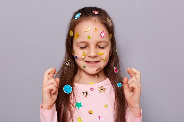 Ritratto di adorabile bambina dai capelli castani sorridente ricoperta di adesivi in posa isolata su sfondo grigio dita incrociate con espressione di speranza occhi chiusi che esprimono desiderio