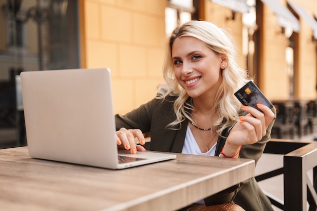 Ritratto di adorabile donna bionda che indossa giacca utilizzando laptop d'argento e tenendo la carta di credito, mentre è seduto in un caffè all'aperto