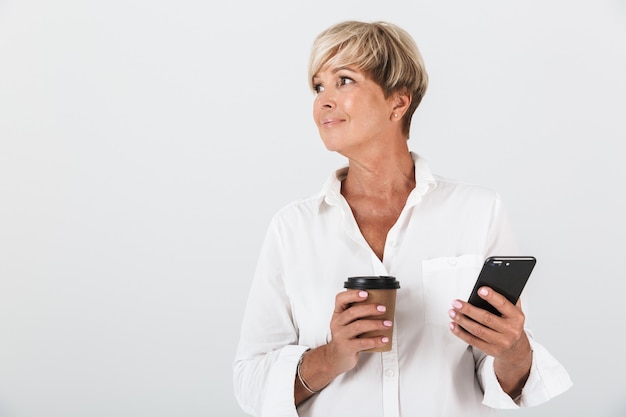 Ritratto di affascinante donna adulta con capelli biondi corti che tiene il cellulare e la tazza di caffè da asporto isolata sul muro bianco in studio