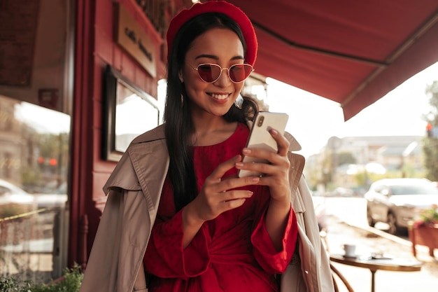 Ritratto di affascinante donna asiatica bruna abbronzata con occhiali da sole rossi abito berretto e trench beige sorride e tiene il telefono all'esterno