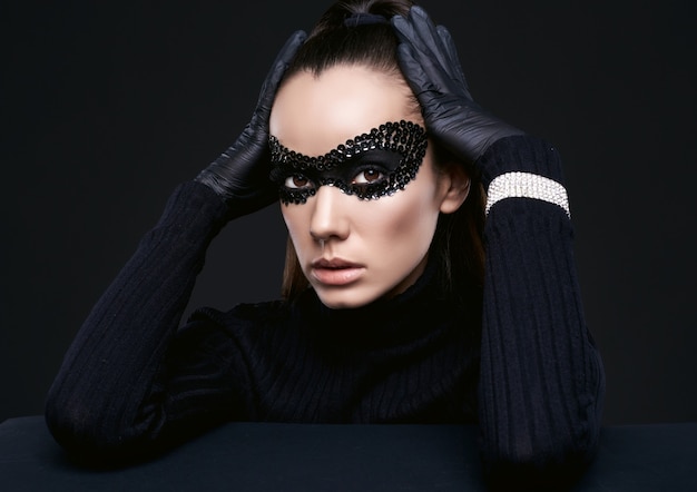 Ritratto di affascinante donna bruna elegante in maglione dolcevita e paillettes maschera in posa sul nero in studio