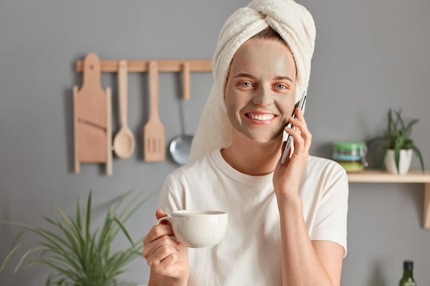 Ritratto di affascinante donna sorridente carina avvolta in un asciugamano che indossa una maglietta bianca in piedi in cucina con in mano una tazza di caffè o tè che parla al telefono con espressione compiaciuta