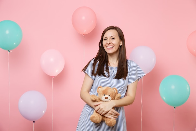 Ritratto di affascinante gioiosa giovane donna in abito blu che tiene e abbraccia un orsacchiotto di peluche su sfondo rosa con mongolfiere colorate. Festa di compleanno persone sincere emozioni concetto.