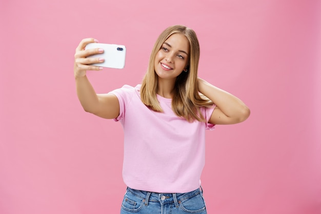 Ritratto di affascinante giovane donna abbronzata in t-shirt toccando i capelli tirando delicatamente la mano con lo smartphone vicino al viso che cattura foto sopra la parete rosa