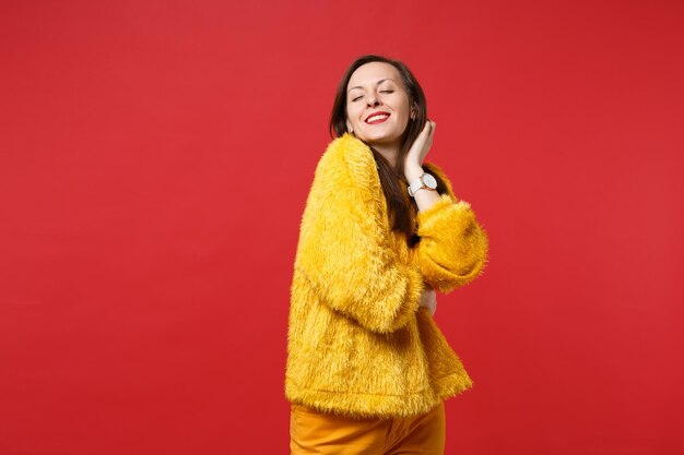 Ritratto di affascinante giovane donna rilassata in maglione di pelliccia gialla che tiene gli occhi chiusi isolati su sfondo rosso brillante della parete in studio. Persone sincere emozioni, concetto di stile di vita. Mock up copia spazio.