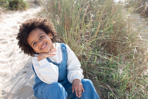 Ritratto di affascinante ragazza afroamericana sulla spiaggia. Modello femminile con capelli ricci in jeans nel complesso seduto sulla sabbia, guardando la fotocamera. Ritratto, concetto di bellezza