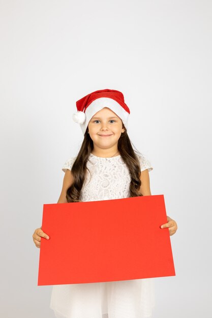 Ritratto di affascinante ragazza dai capelli lunghi in abito bianco e cappello di babbo natale con striscione bianco rosso iso...