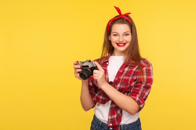 Ritratto di affascinante ragazza pinup felice in camicia a scacchi e fascia per capelli che tiene una macchina fotografica vecchio stile e sorride alla macchina fotografica, scattando foto godendosi l'hobby. stile retrò anni '50, girato in studio al coperto isolato