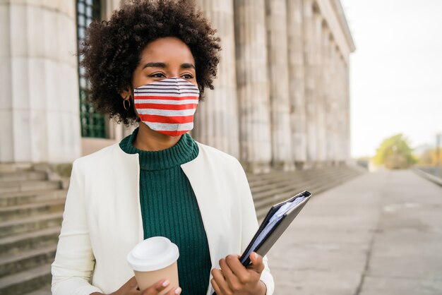 Ritratto di afro imprenditrice con maschera protettiva tenendo una tazza di caffè e appunti