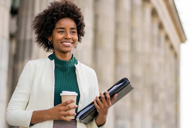 Ritratto di afro imprenditrice tenendo una tazza di caffè e appunti mentre si cammina all'aperto in strada. Business e concetto urbano.