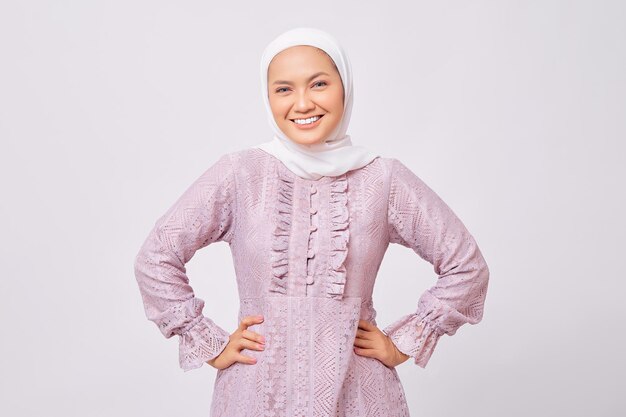 Ritratto di allegra bella giovane donna musulmana asiatica che indossa hijab e abito viola che si tiene per mano sulla vita guardando la fotocamera isolata su sfondo bianco studio