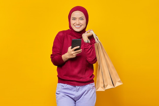 Ritratto di allegra donna asiatica che tiene telefono e borsa della spesa su sfondo giallo