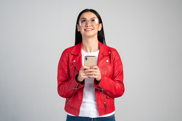 Ritratto di allegra donna bruna alzando lo sguardo mentre si utilizza il cellulare e sorridente. Femmina che legge un messaggio di buone notizie, godendo dell'applicazione mobile. Colpo dello studio dell'interno isolato su fondo grigio