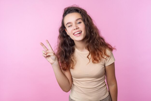 Ritratto di allegra vivida ragazza adolescente con lunghi capelli ricci castani che indossa abiti beige in stile casual che strizzano l'occhio alla fotocamera e mostrano il gesto di vittoria. studio indoor girato isolato su sfondo rosa