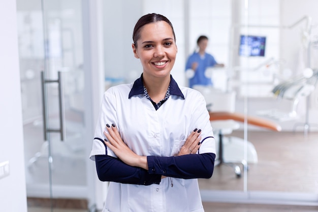 Ritratto di allegro dentista in reception dentale con le braccia incrociate guardando la telecamera indossando l'uniforme. Dentista sorridente felice nel suo ufficio ortodontico.