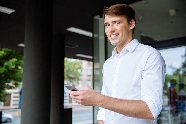 Ritratto di allegro giovane imprenditore utilizza lo smartphone e sorridente vicino al centro business