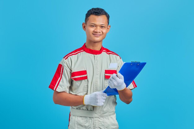 Ritratto di allegro giovane meccanico asiatico che tiene appunti isolati su sfondo blu