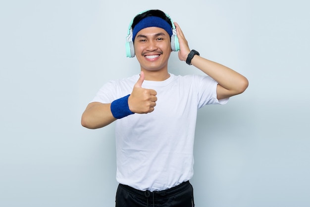 Ritratto di allegro giovane sportivo asiatico in fascia blu e maglietta bianca di abbigliamento sportivo mentre si ascolta la musica preferita con le cuffie che mostrano il pollice in alto gesto isolato su sfondo bianco