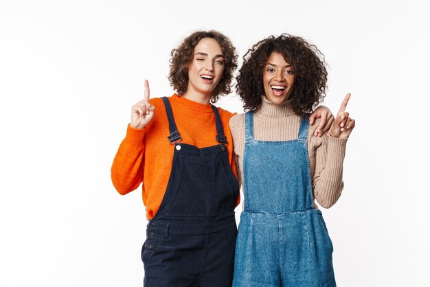 Ritratto di allegro ottimista due amiche multirazziali in tute di jeans isolate sul muro bianco che punta a copyspace.