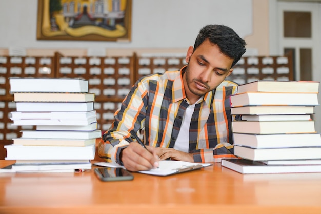 Ritratto di allegro studente indiano internazionale maschile con accessori per l'apprendimento dello zaino in piedi vicino agli scaffali della biblioteca universitaria o del negozio di libri durante la pausa tra le lezioni Concetto di educazione